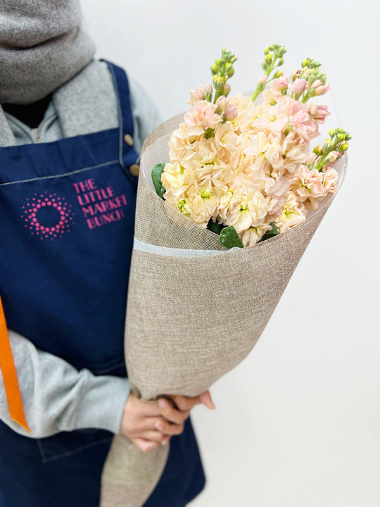 A person holding a medium-sized bouquet of assorted pink and white stock flowers, wrapped in beige paper with a visible brand logo on the attire.