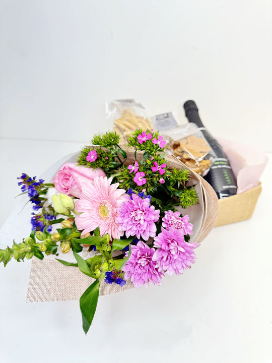 
                  
                    A beautifully arranged gift set featuring a bouquet of pink and purple flowers including gerbera, roses, and chrysanthemums, paired with a bottle of Maschio Dei Cavalieri Treviso Prosecco and a selection of packaged gourmet snacks in a Small hessian box. Shot by The Little Market Bunch, Melbourne.
                  
                