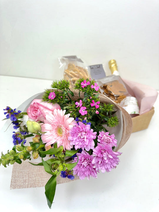 
                  
                    A beautifully arranged gift set featuring a bouquet of pink and purple flowers including gerbera, roses, and chrysanthemums, paired with a bottle of Pink Grapefruit Sparkling and a selection of packaged gourmet snacks in a Small hessian box. Shot by The Little Market Bunch, Melbourne.
                  
                