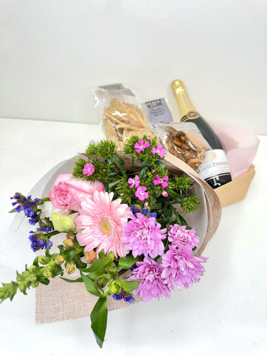 
                  
                    A beautifully arranged gift set featuring a bouquet of pink and purple flowers including gerbera, roses, and chrysanthemums, paired with a bottle of Louis Perdrier sparkling wine and a selection of packaged gourmet snacks in a Small hessian box. Shot by The Little Market Bunch, Melbourne.
                  
                