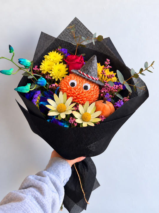 A hand holding a unique decorative bouquet wrapped in black tissue paper. The bouquet features a central orange fuzzy owl plush wearing a pointed hat, surrounded by yellow chrysanthemums, red roses, purple and blue flowers, and small orange pumpkins, with green eucalyptus stems, against a plain white background.