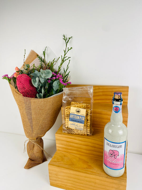 Bouquet of flowers, bag of Artisan Pizza, and bottle of Delirium Tremens on a wooden stand.