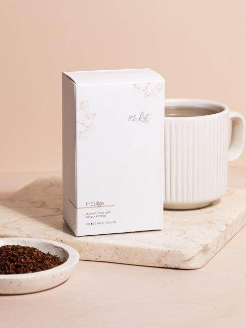 White tea box labeled 'P.S. Tea' next to a white mug and bowl of tea leaves on a beige surface