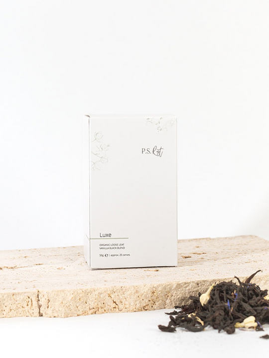 
                  
                    White box labeled 'P.S. Tea' on a wooden surface with loose tea leaves.
                  
                