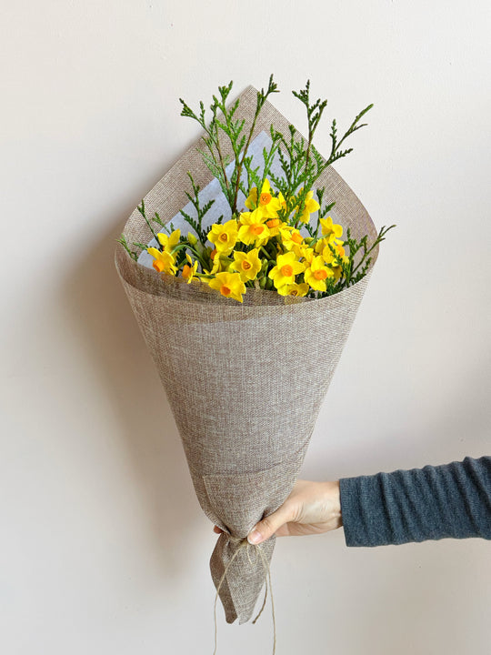 
                  
                    A bouquet of yellow flowers with green stems wrapped in a textured beige burlap cone, held by a hand wearing a dark blue long-sleeve shirt against a plain off-white wall.
                  
                
