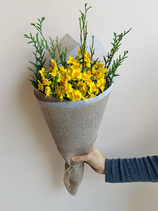 
                  
                    A hand wrapped in a blue long-sleeve shirt holds a bouquet of yellow flowers and green foliage wrapped in textured beige paper against a plain light background.
                  
                