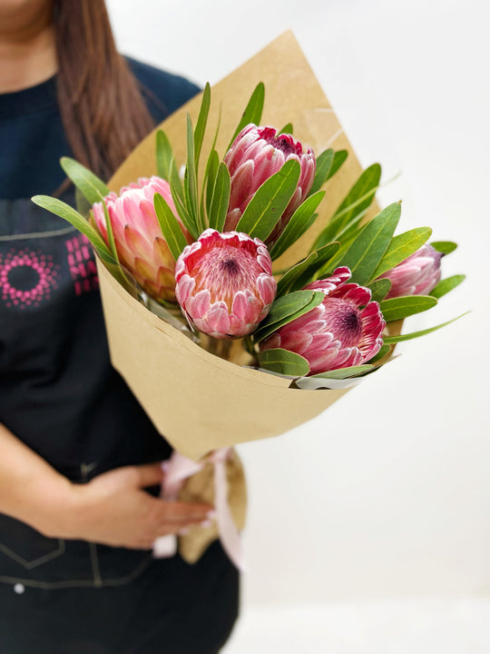 A person holding a bouquet of pink and red Protea flowers wrapped in beige paper.