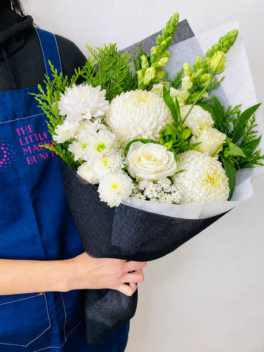 Person holding a bouquet of white flowers with greenery, wearing a blue apron with 'The Little Market Bunch' branding.